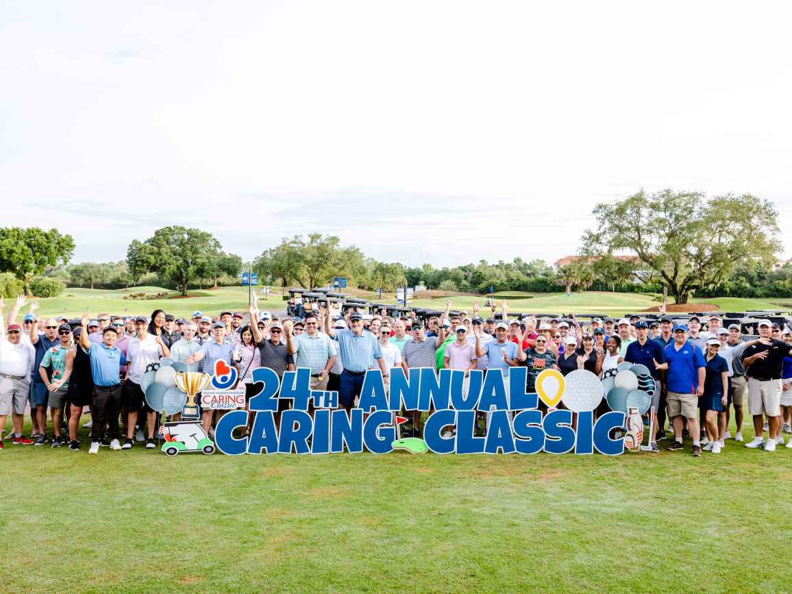A group of people happily standing behind the Caring Classic golf event sign